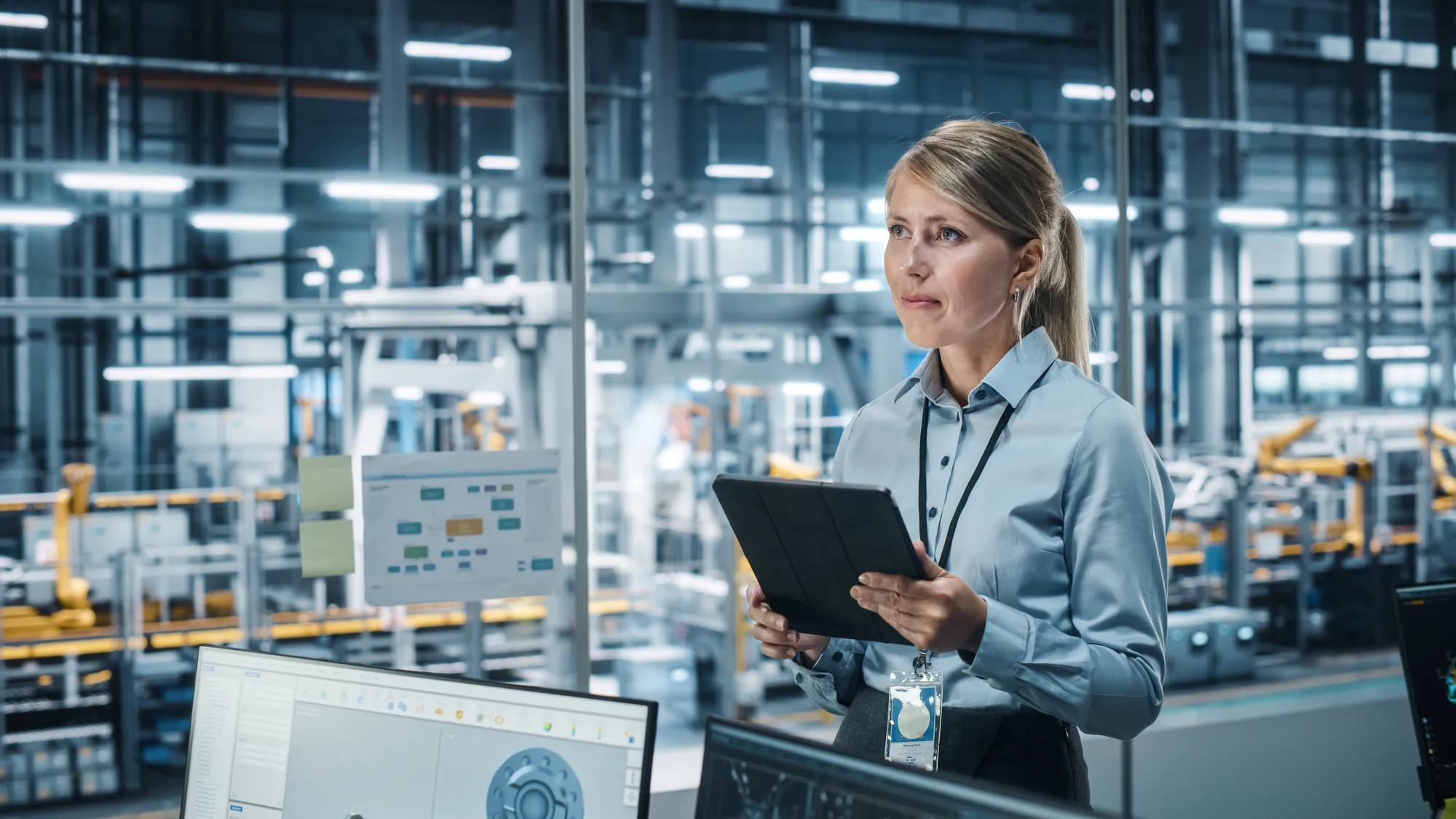 Car Factory Office Concept: Portrait of Successful Female Chief Automotive Engineer Using Tablet Computer to Design and Optimize Automated Robot Arm Assembly Line Manufacturing Electric Vehicles