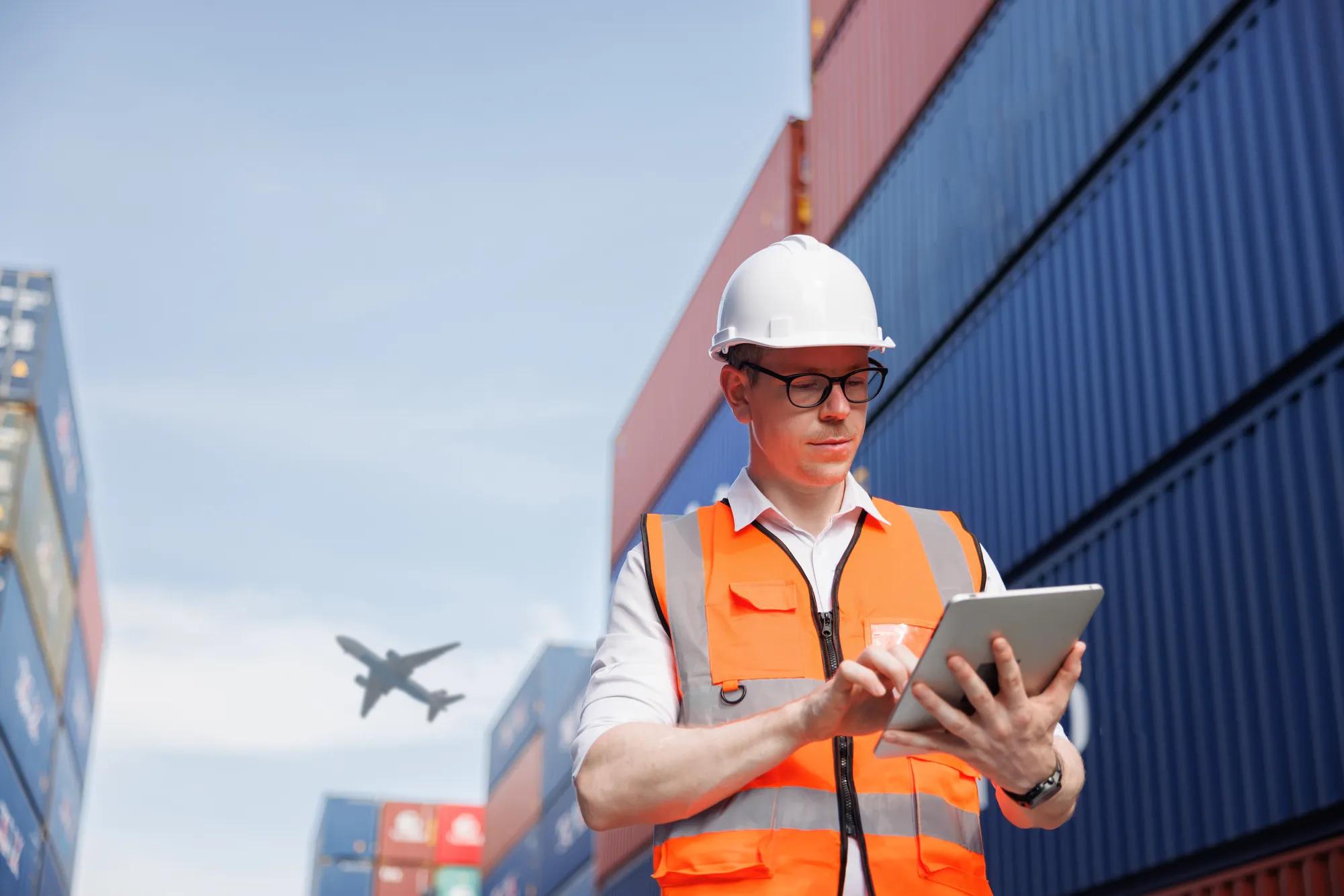 the logistic business owner is wearing a white safety helmet and using the digital tablet in a Cargo freight ship for import export container yard with airplane on sky background . planing engineer.